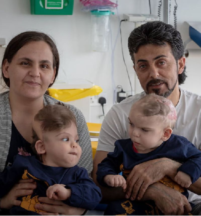 The parents of Yigit and Derman hold their sons post-surgery, and look at the camera. Hospital equipment is in the background.
