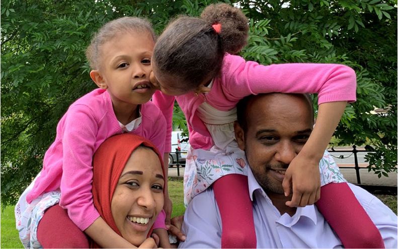 Rital and Ritag sit on their parent's shoulders. One kisses the other, while their parents smile for the camera.