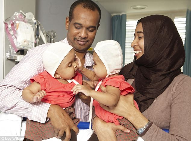 Rital and Ritag's parents hold their daughters post-operation. The babies interact with each other.